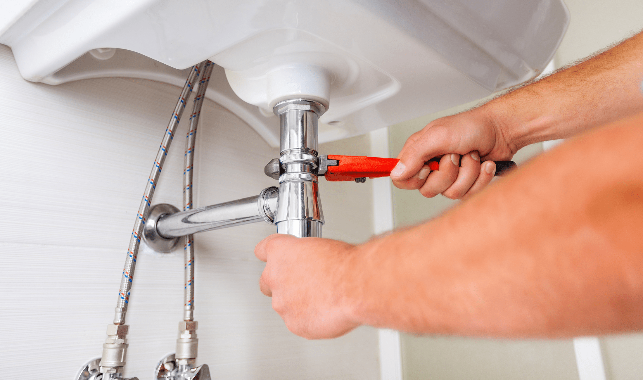 A pair of hands using an adjustable wrench to tighten a joint in a sink's pipes.
