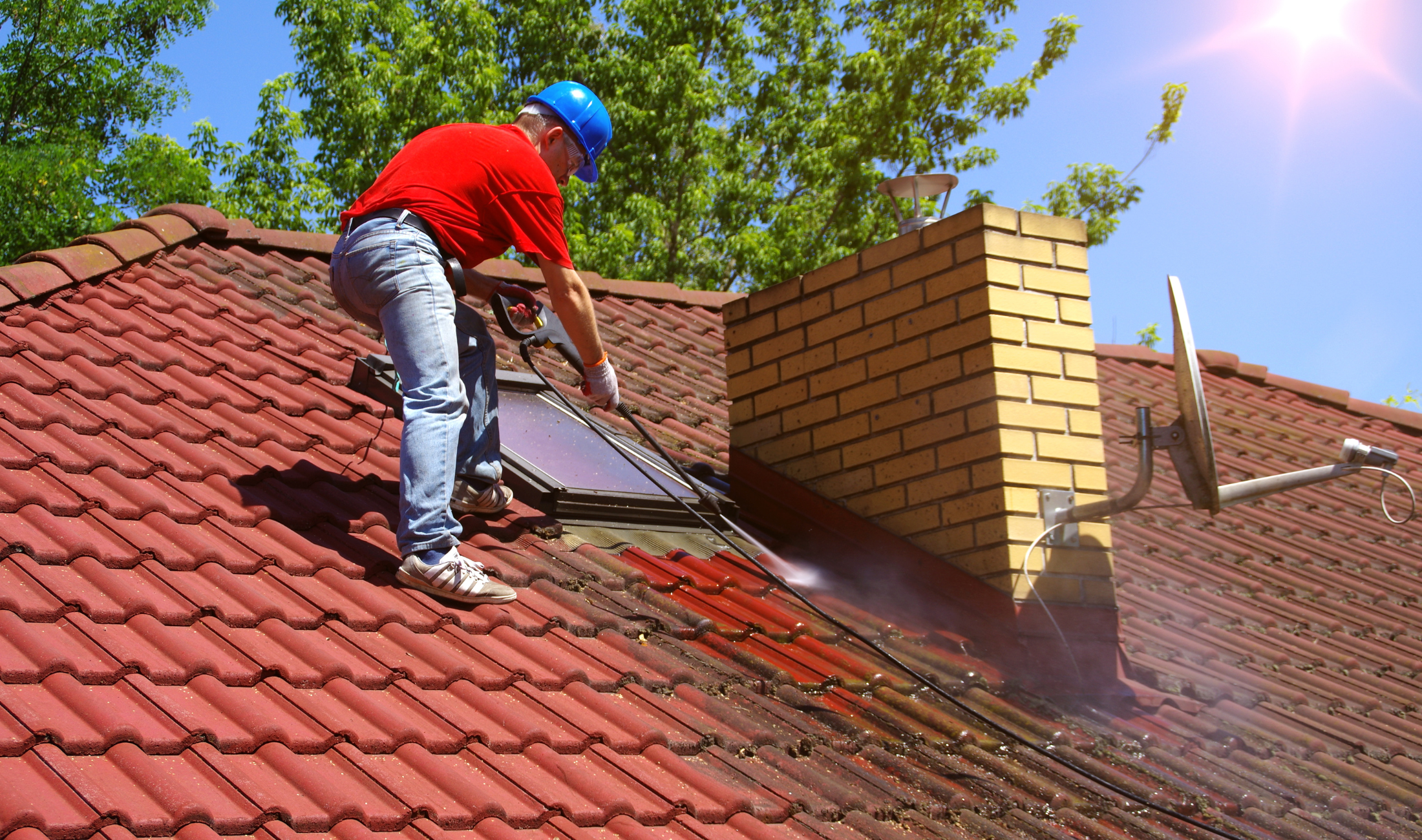 A man standing on a roof pressure washing the shingles.