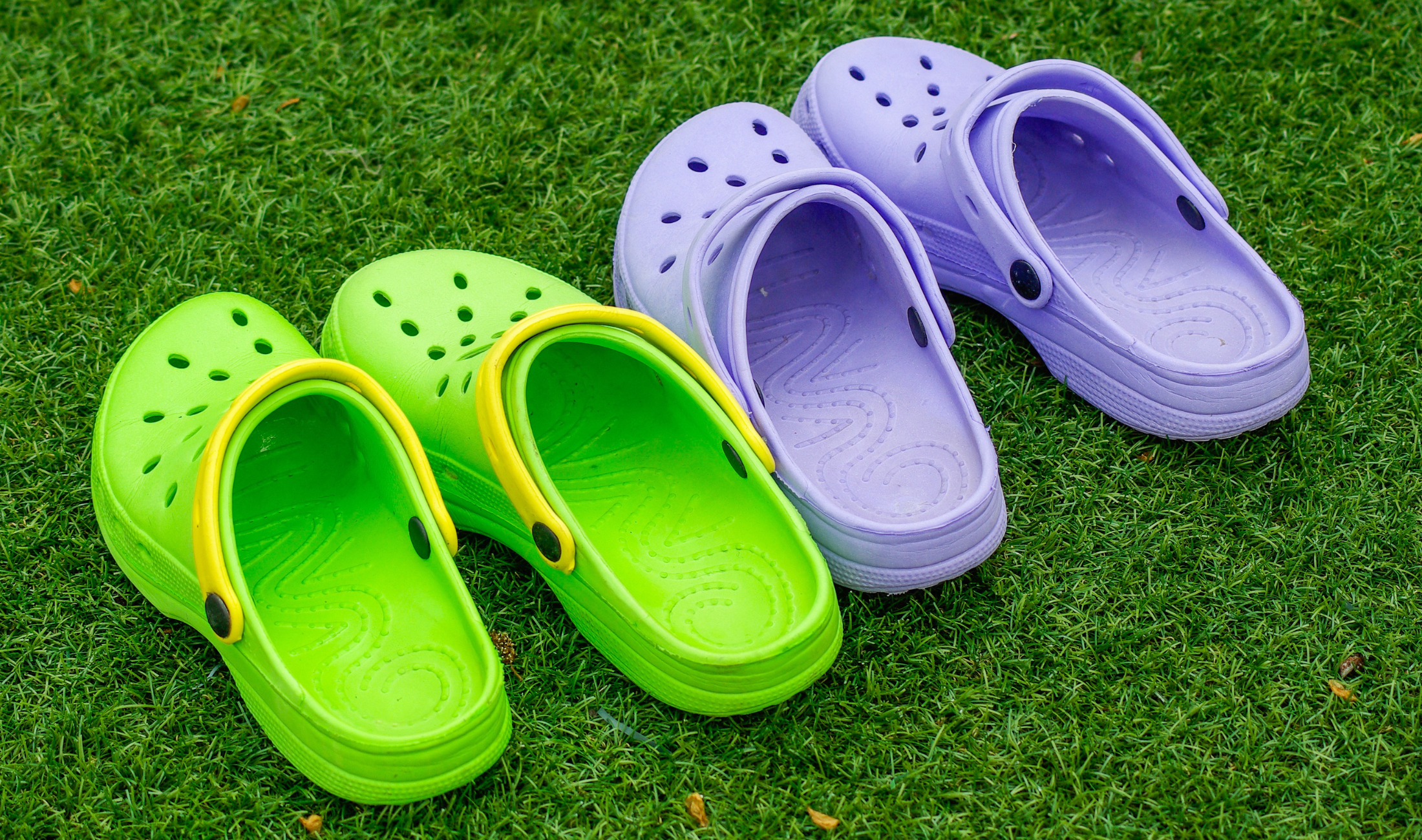 A pair of rubber crocks on grass, one green pair and one purple pair.
