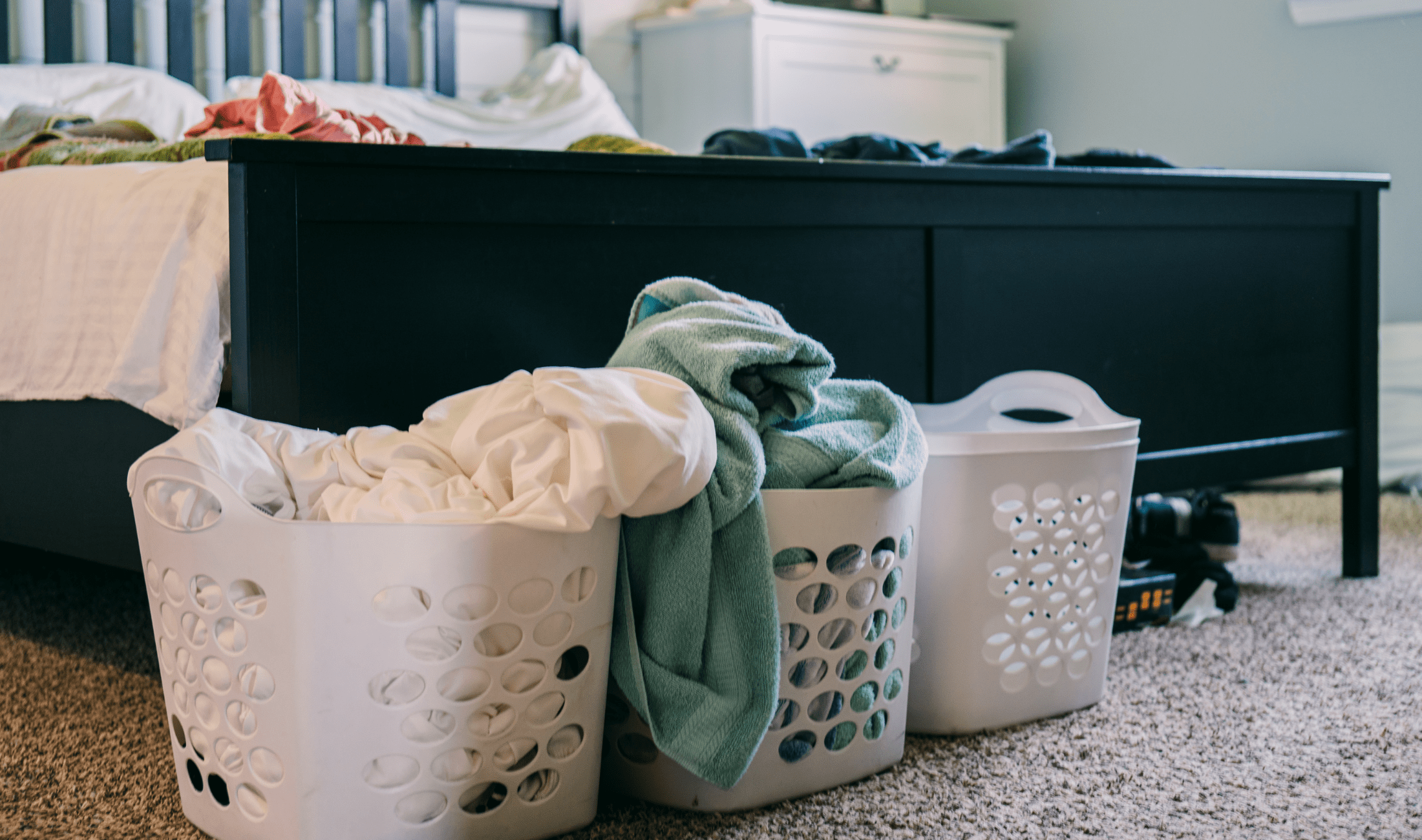 Three small laundry hampers in front of a bed, some with clothes in them.