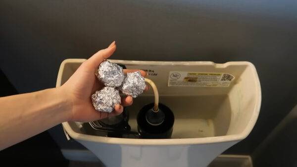 A hand holding three bowls of aluminum foil above an open toilet tank.