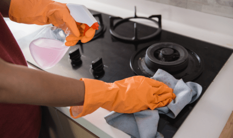 A pair of gloved hands washing a stove with a spray bottle of cleaner.