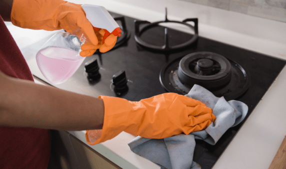 A pair of gloved hands washing a stove with a spray bottle of cleaner.