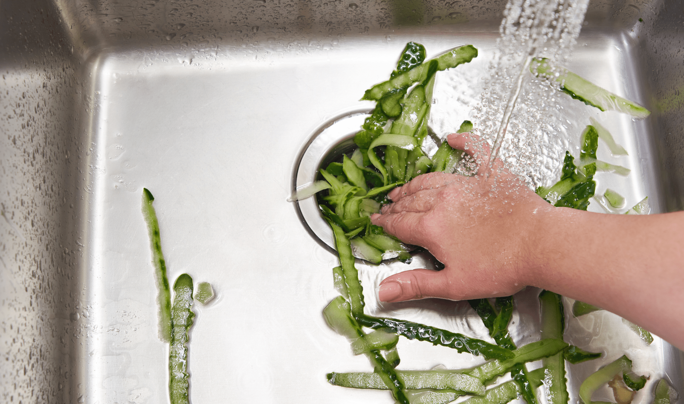A woman's hand pushing cucumber peels down a sink's garbage disposal.