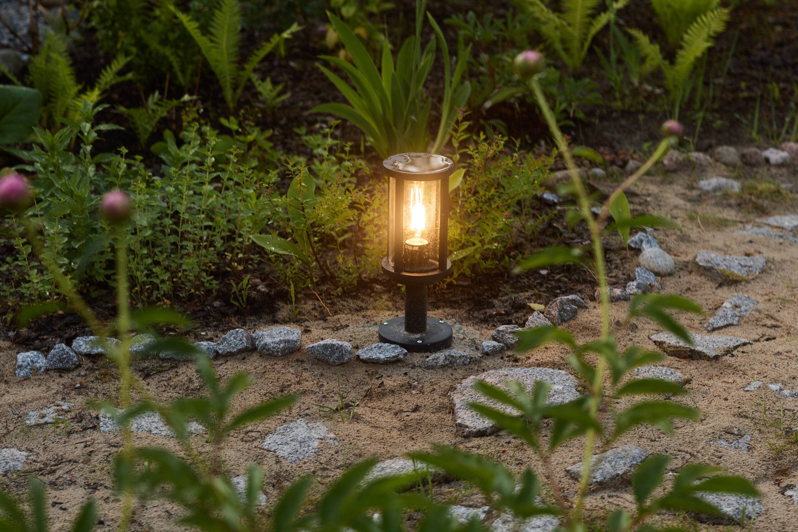 A softly glowing garden lantern is beautifully surrounded by lush plants and ornamental stones in a tranquil setting