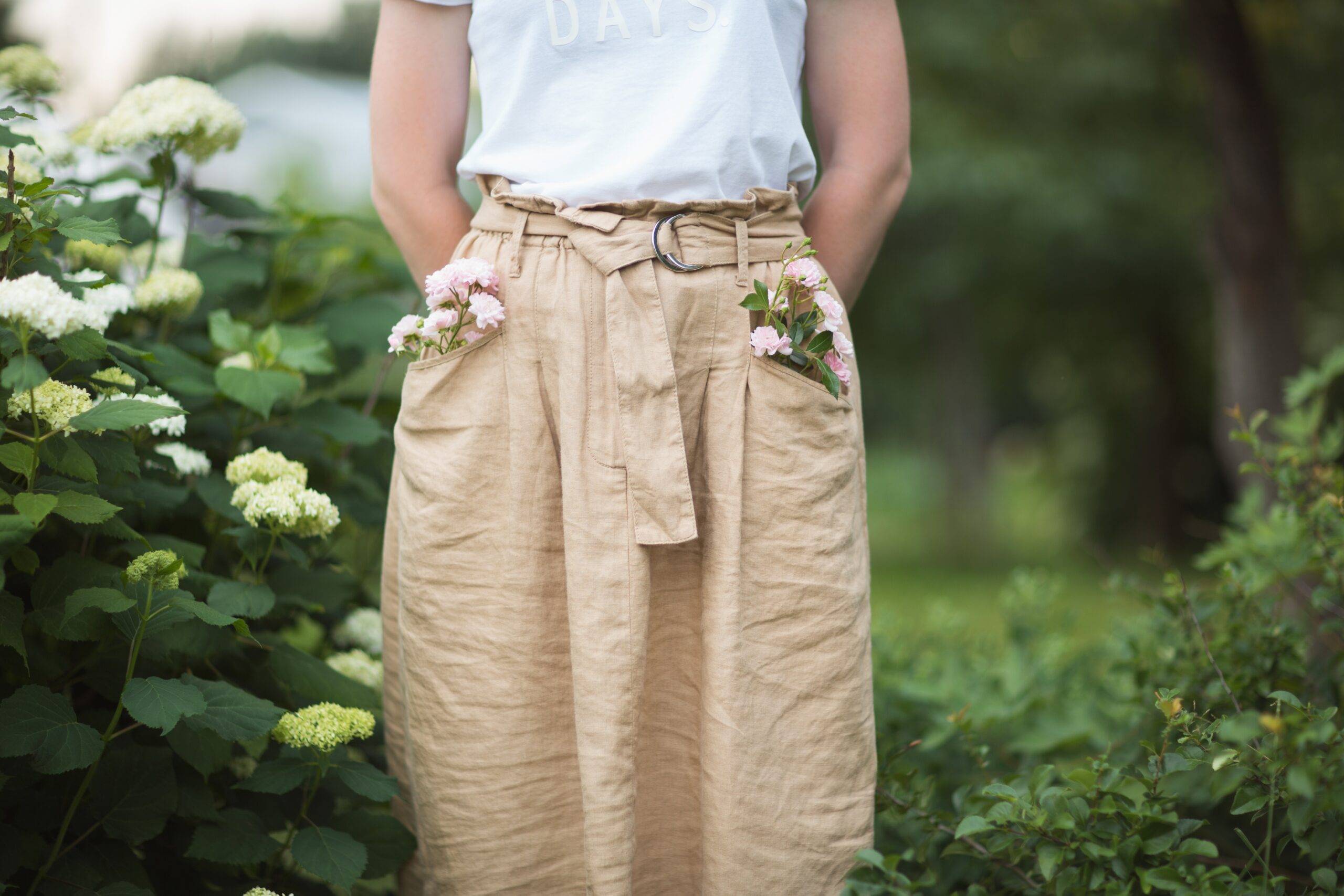 Woman in light brown linen pants and a white shirt, photographed from the waist to the calves, hands in pockets, flowers also in pockets.