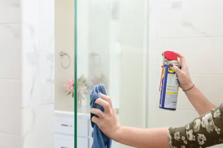 Человек, держащий банку с WD-40 в одной руке, нацеленный на его стеклянную душевую дверь, тряпку в другой руке, вытирая ее.