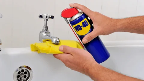 A person holding a can of WD-40 up to a sink faucet, a rag in the other hand.
