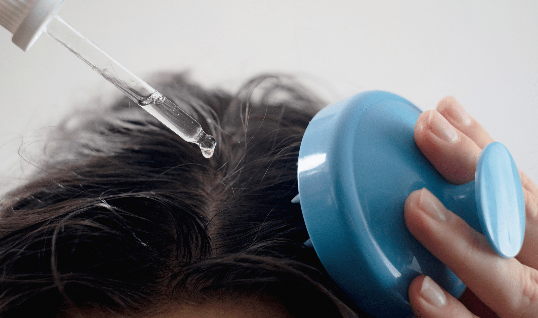 A person applying oil to their scalp with a dropper and a massager.