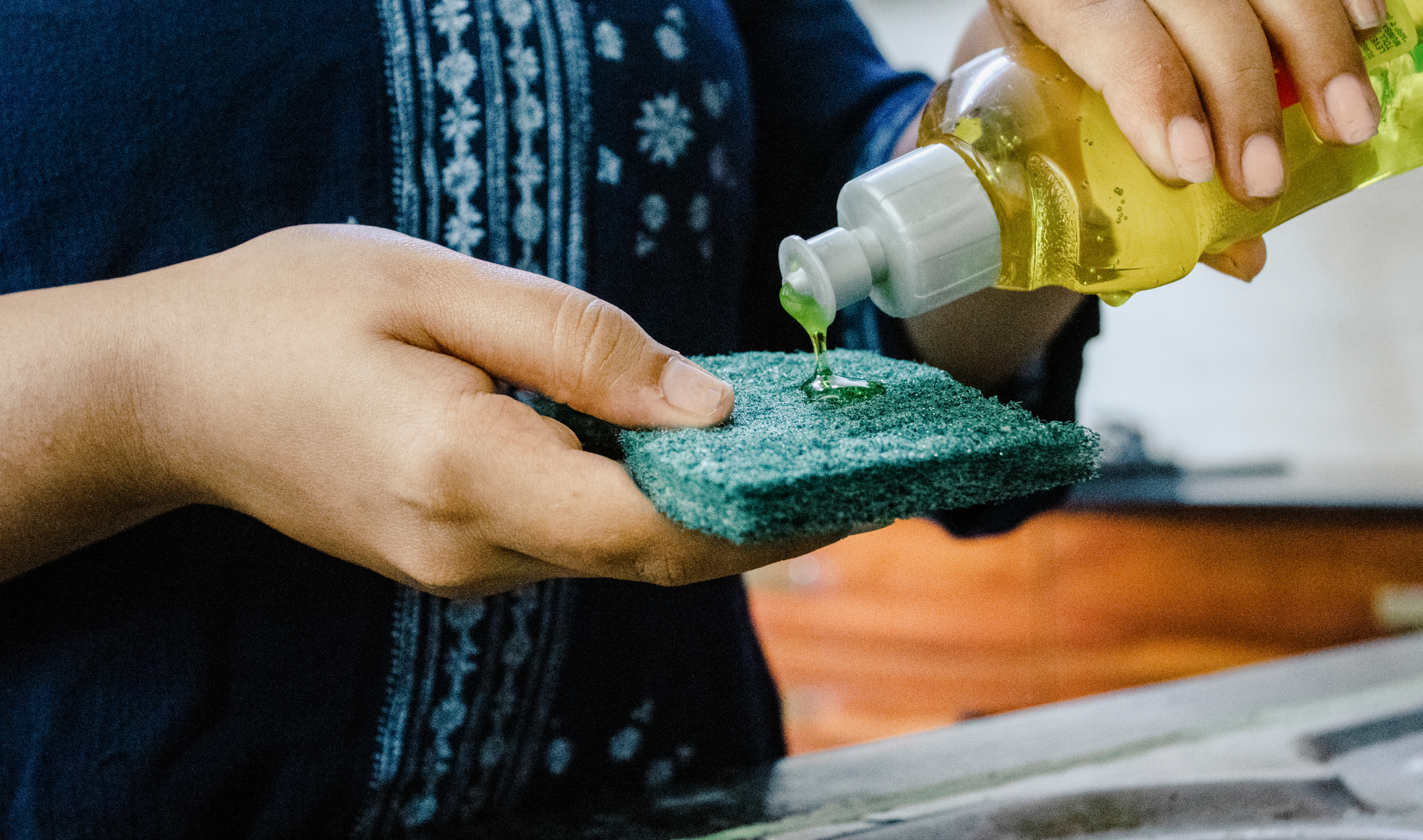 A person holding a dish sponge, squeeing dish soap onto it.