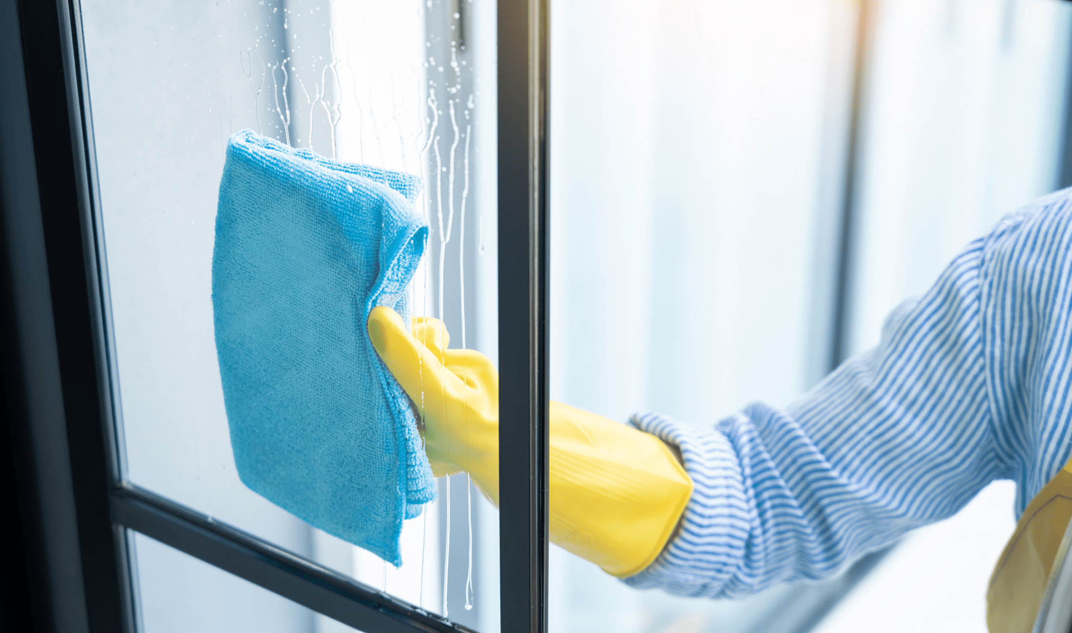 A person in yellow rubber gloves using a blue microfiber cloth on a window that's been sprayed with cleaner.