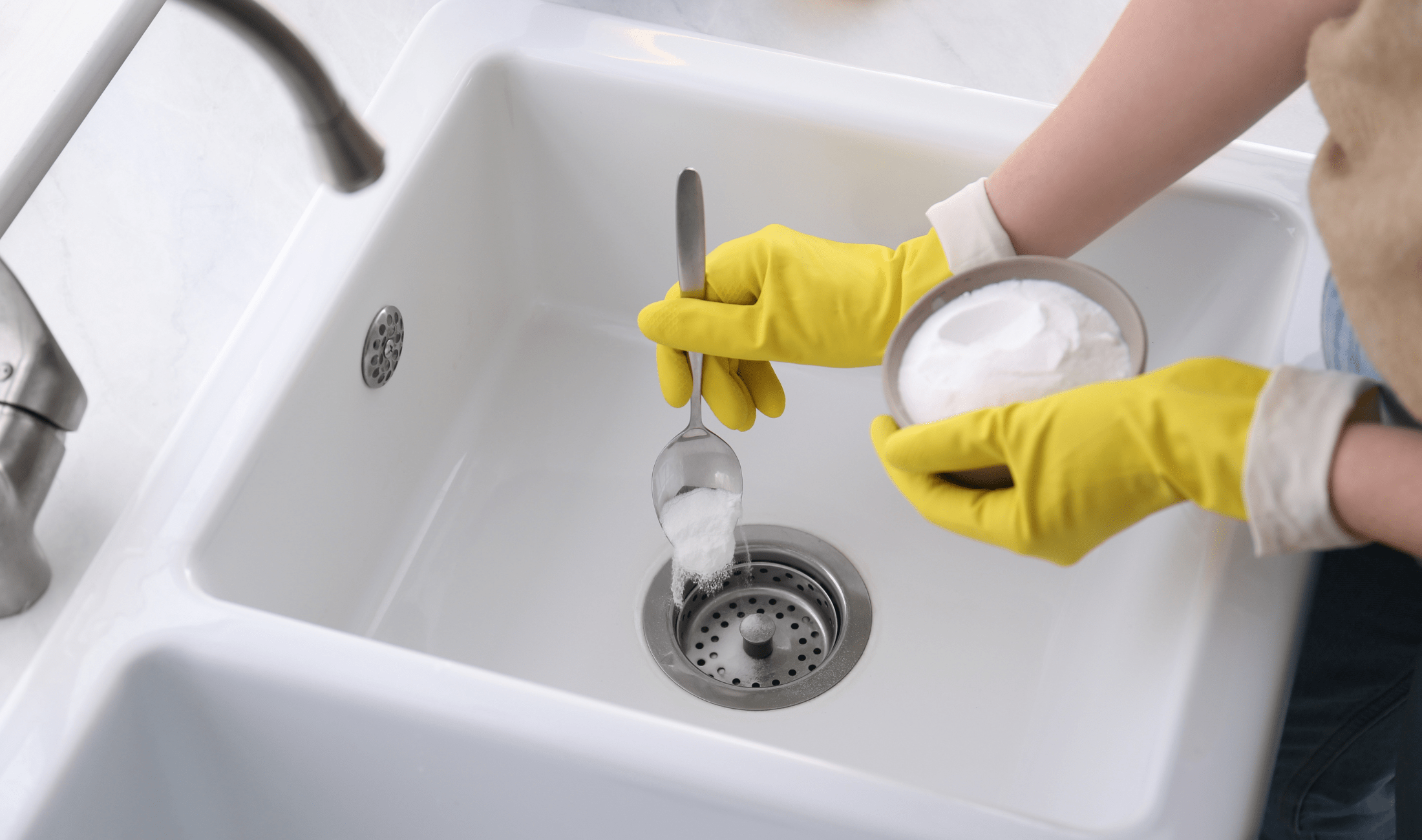 A person wearing yellow gloves sprinkling some baking soda into a kitchen sink drain.