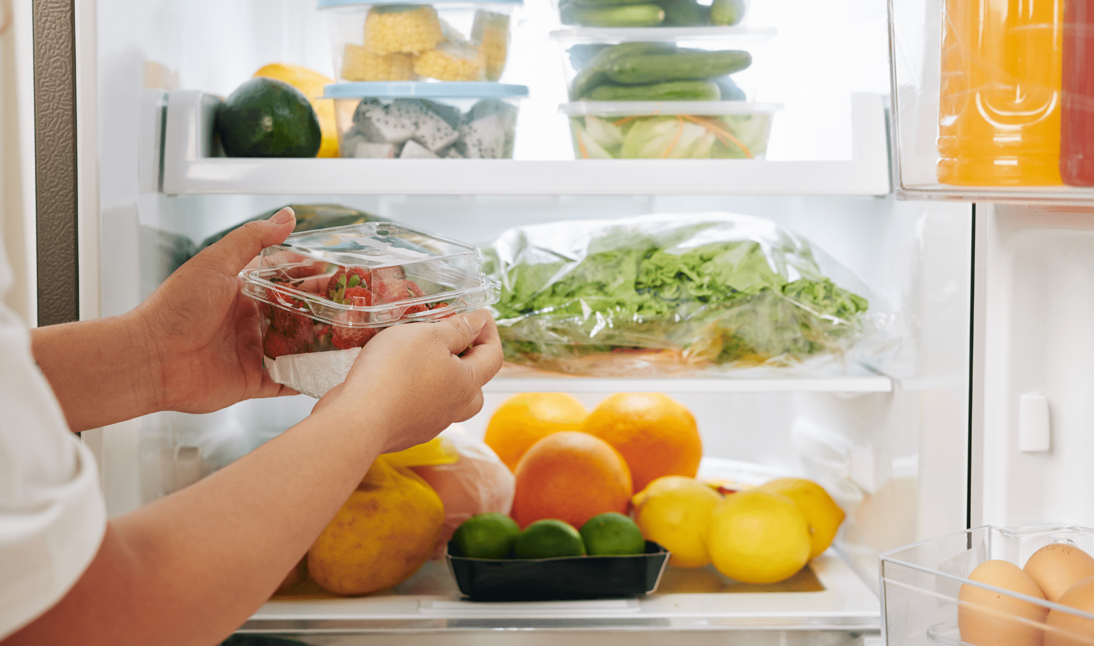 A person putting a container of strawberries into a fridge filled with fresh produce.