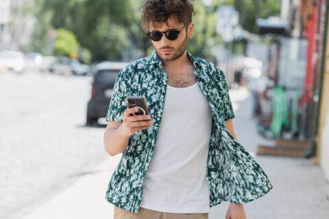 Brunette man in sunglasses using smartphone while walking on urban street