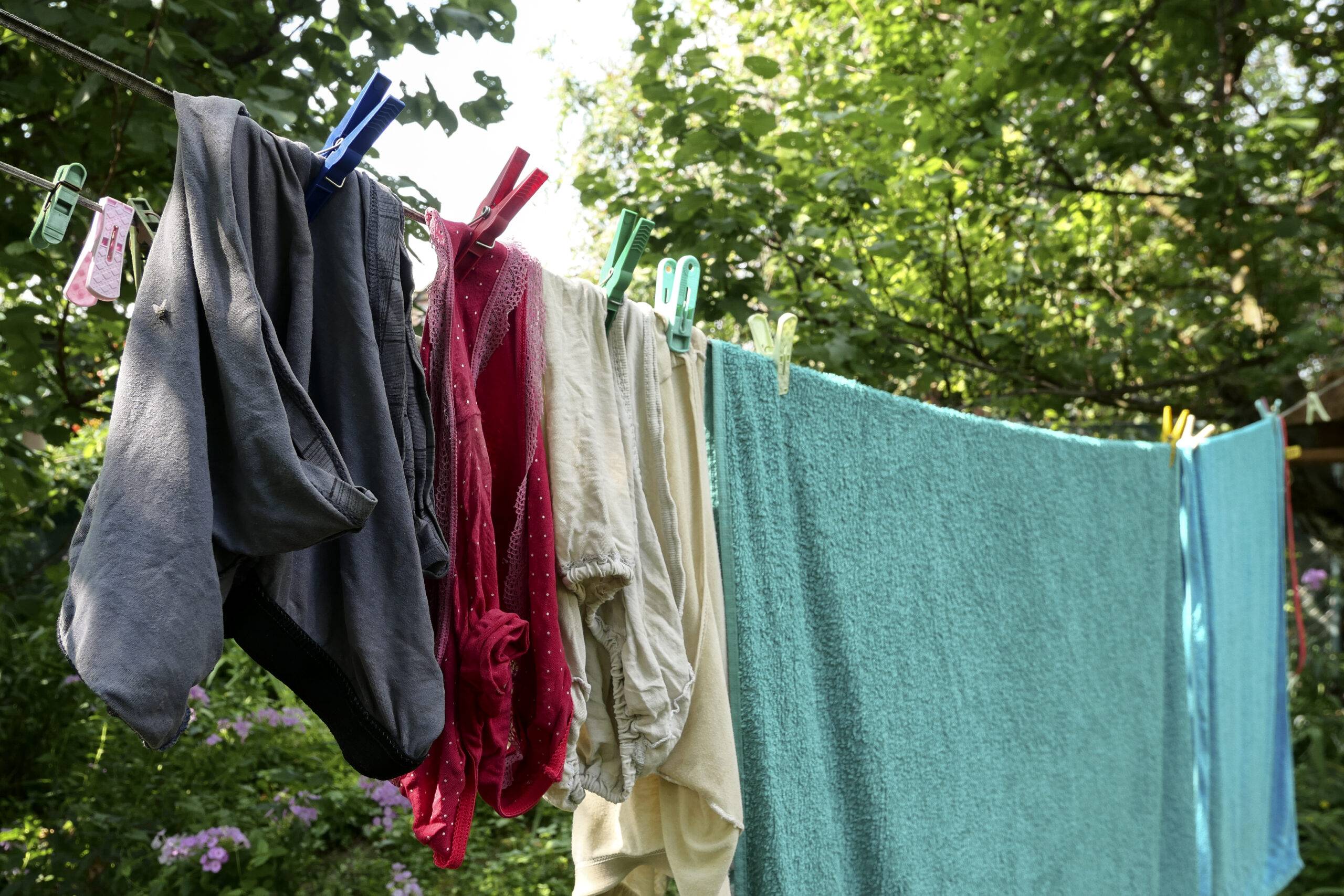 Different washed clothes is dried on rope in backyard or garden. Drying linen with multicolored clothespins. Summer sunny day. Close-up. Outdoors.