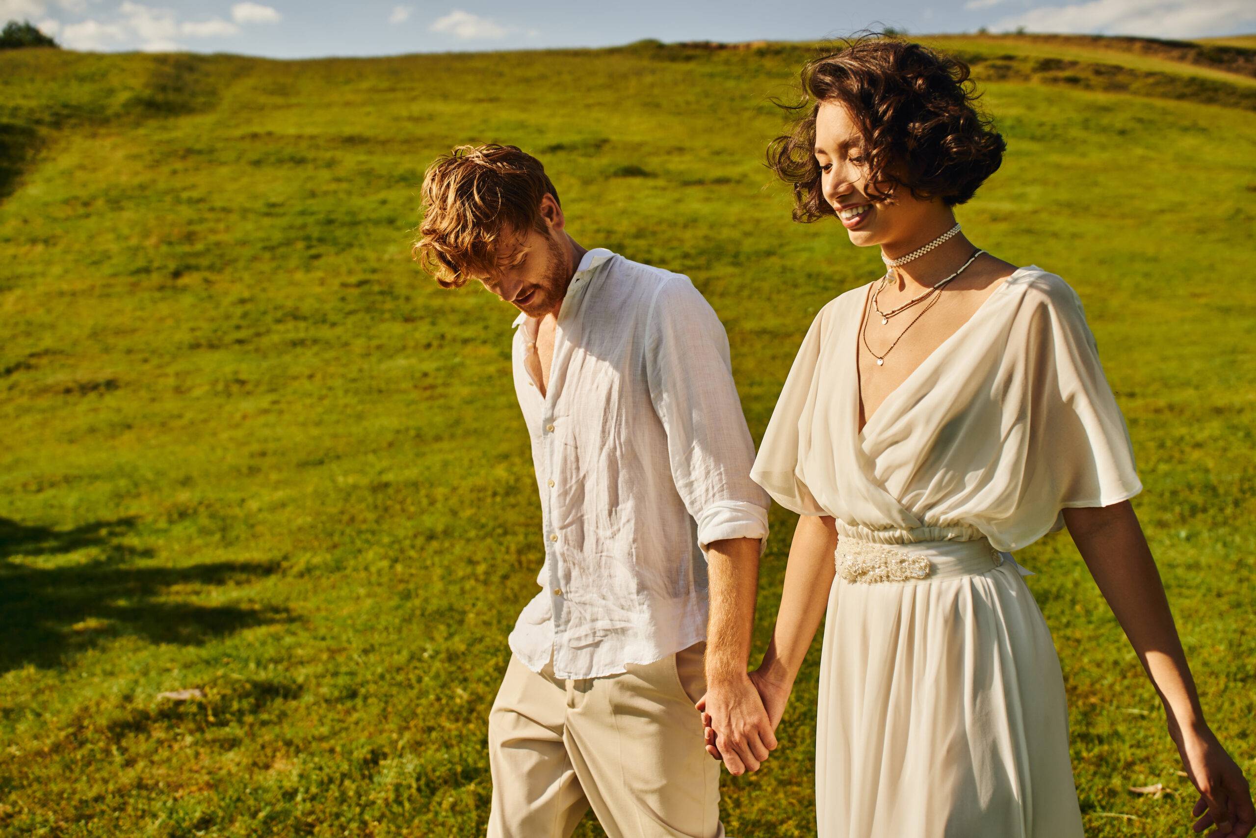 wedding in countryside, happy interracial couple holding hands in green field, just married