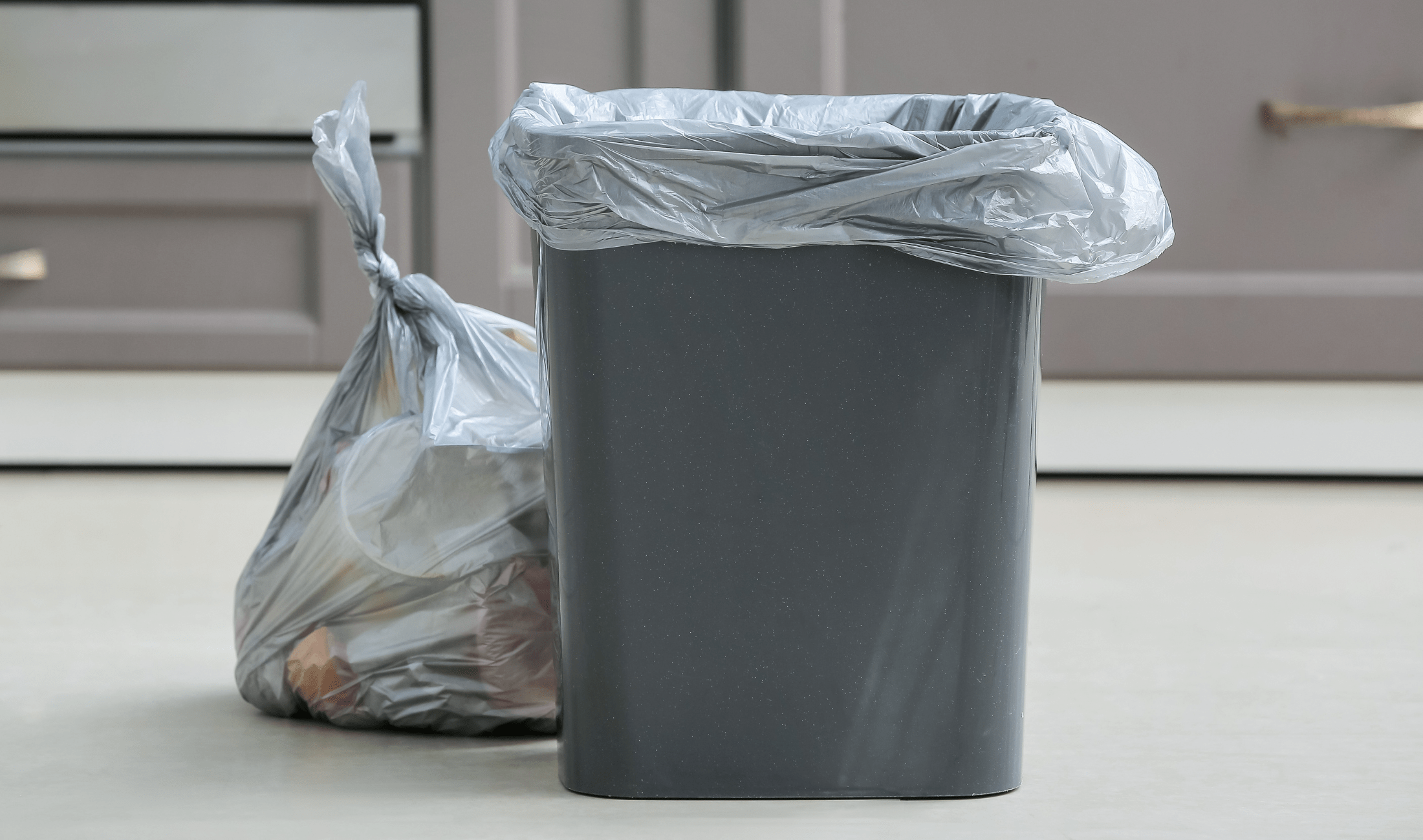 A small, grey, plastic garbage bin, a tied bag next to it.