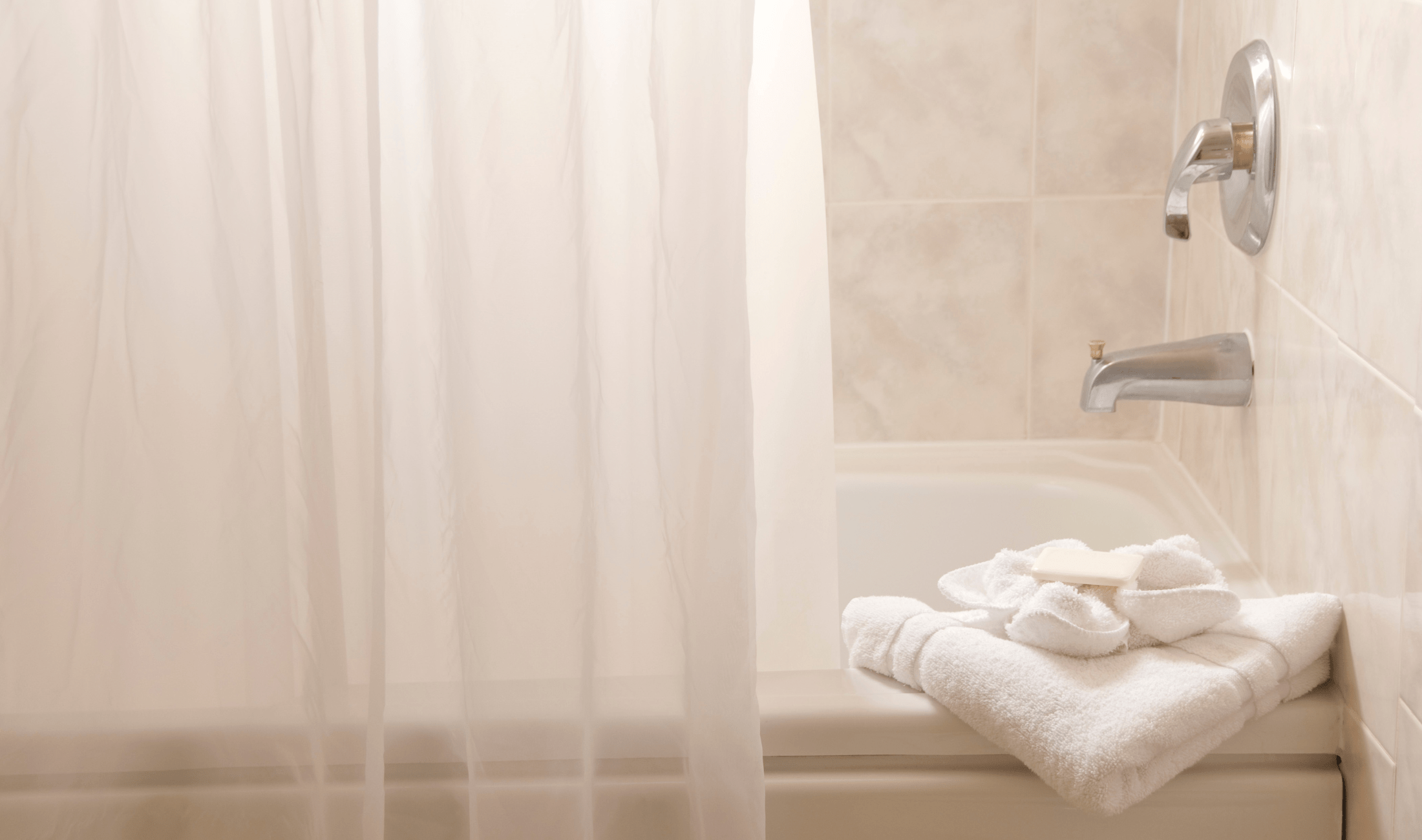 A low photo of the edge of a bath, the shower curtain mostly closed, rolled towels on the corner edge of the tub.