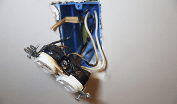 An outlet hganging out of the wall, having clearly sparked or caught fire.