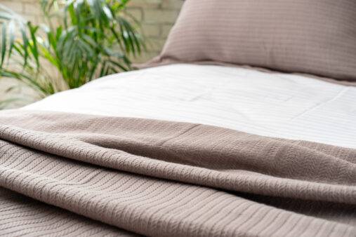 A soft, brown knit blanket lies on a made bed, with white sheets and a pillow visible in the background.