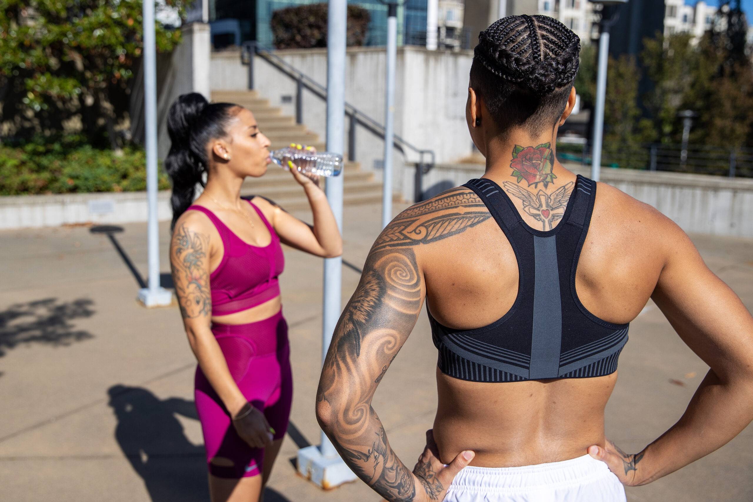 Two women standing outside in athletic wear, one in profile drinking some water, the other facing away from the camera, looking at the other woman.