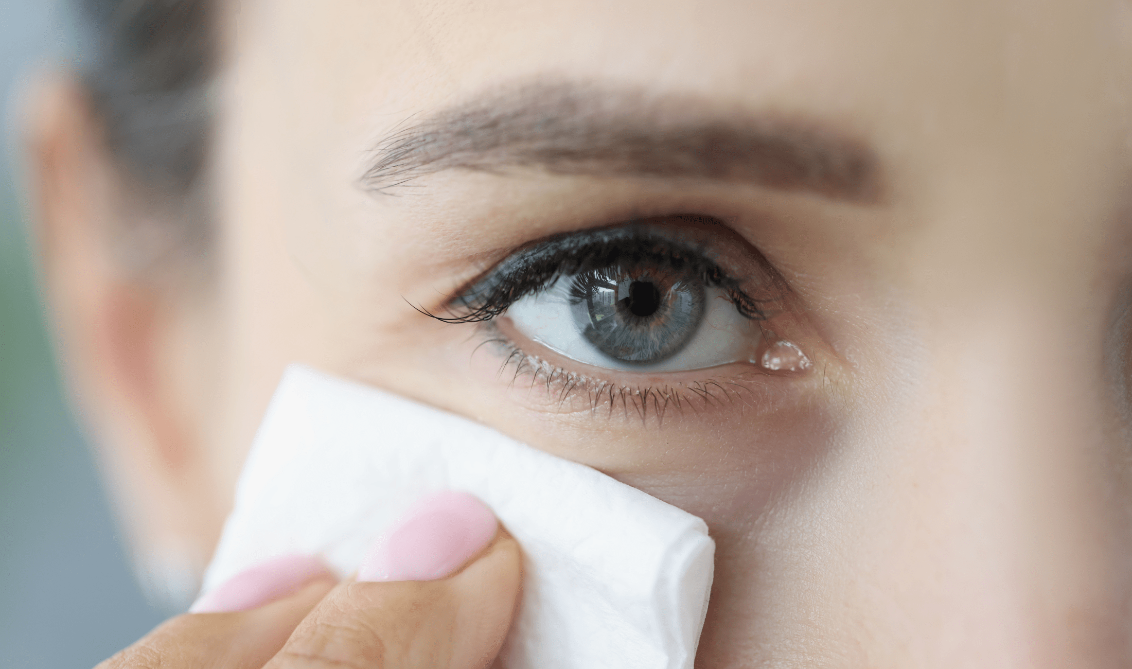 A closeup of a woman's eye, her hand holding a wipe up to her cheek just below.