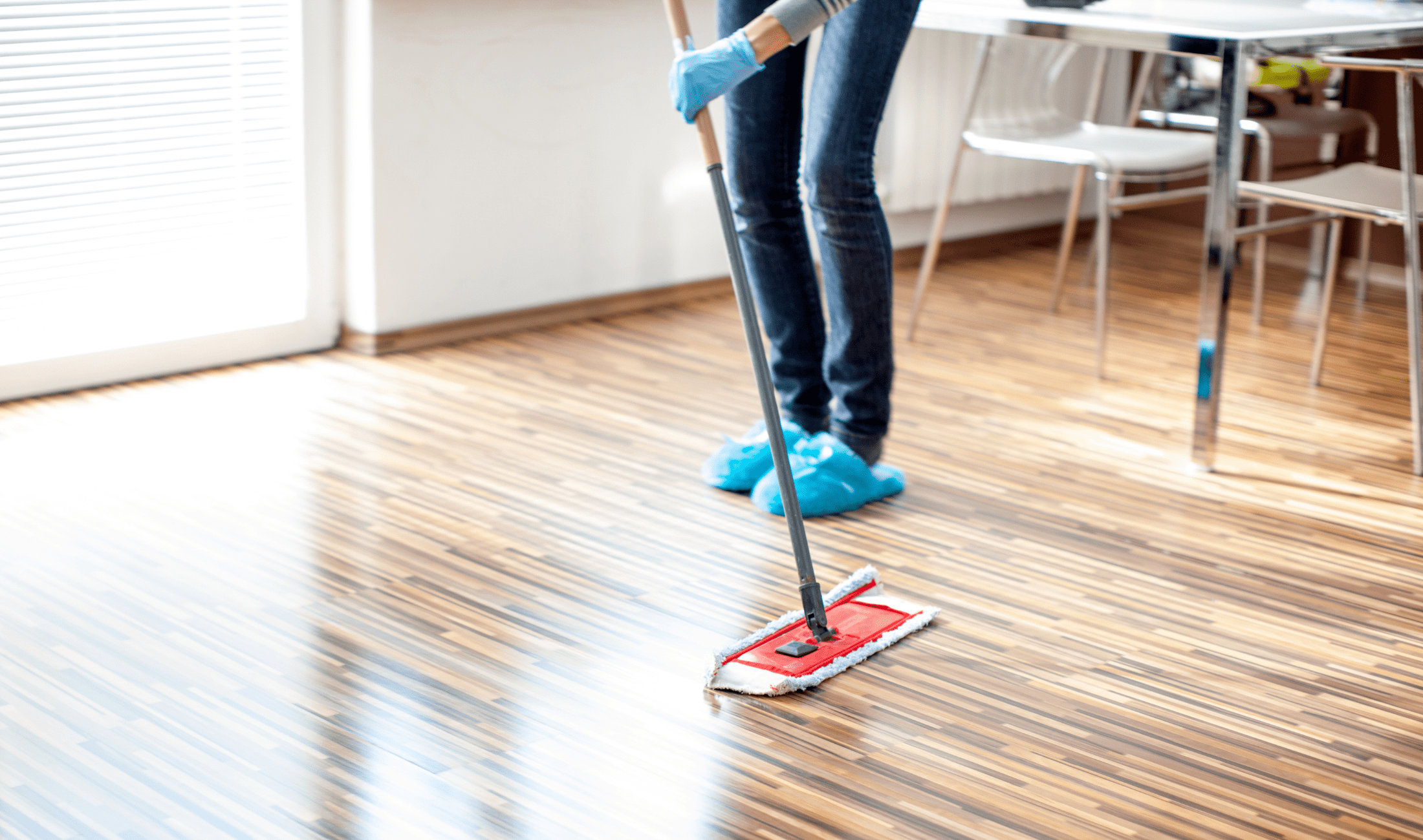 A person in blue slippers and rubber gloves mopping a hardwood floor.