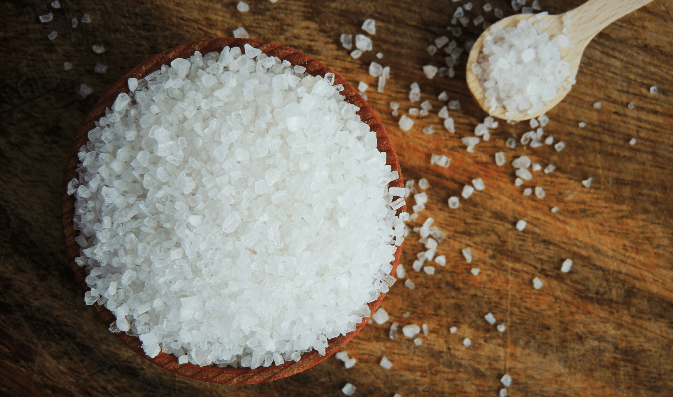 A bowl of epsom salt atop a wooden table, some salt spilling over the side.