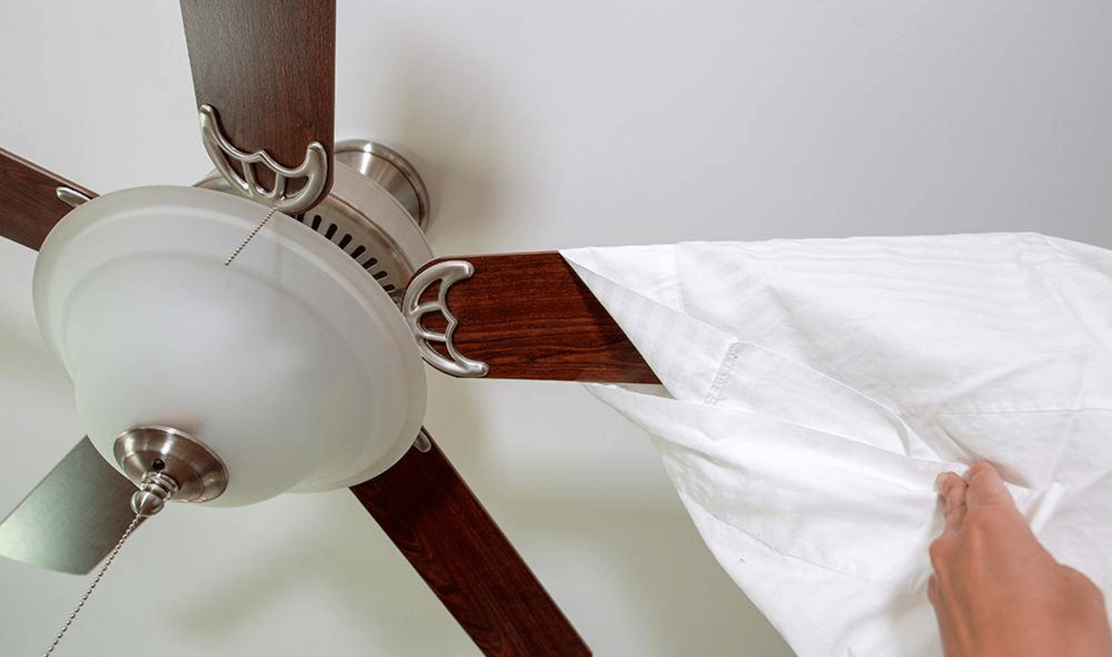 A person using a pillowcase to clean an ceiling fan blade.