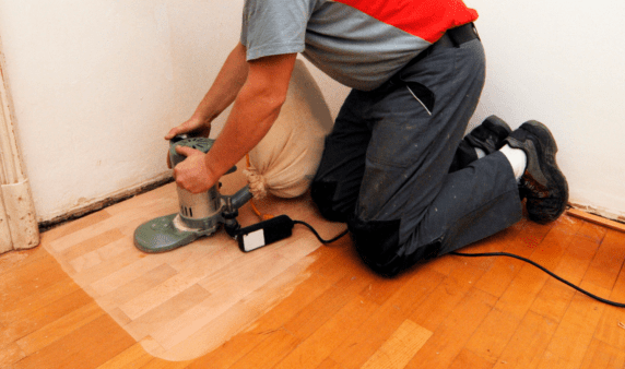 A man kneeling on the ground, sanding a section of a hardwood floor.