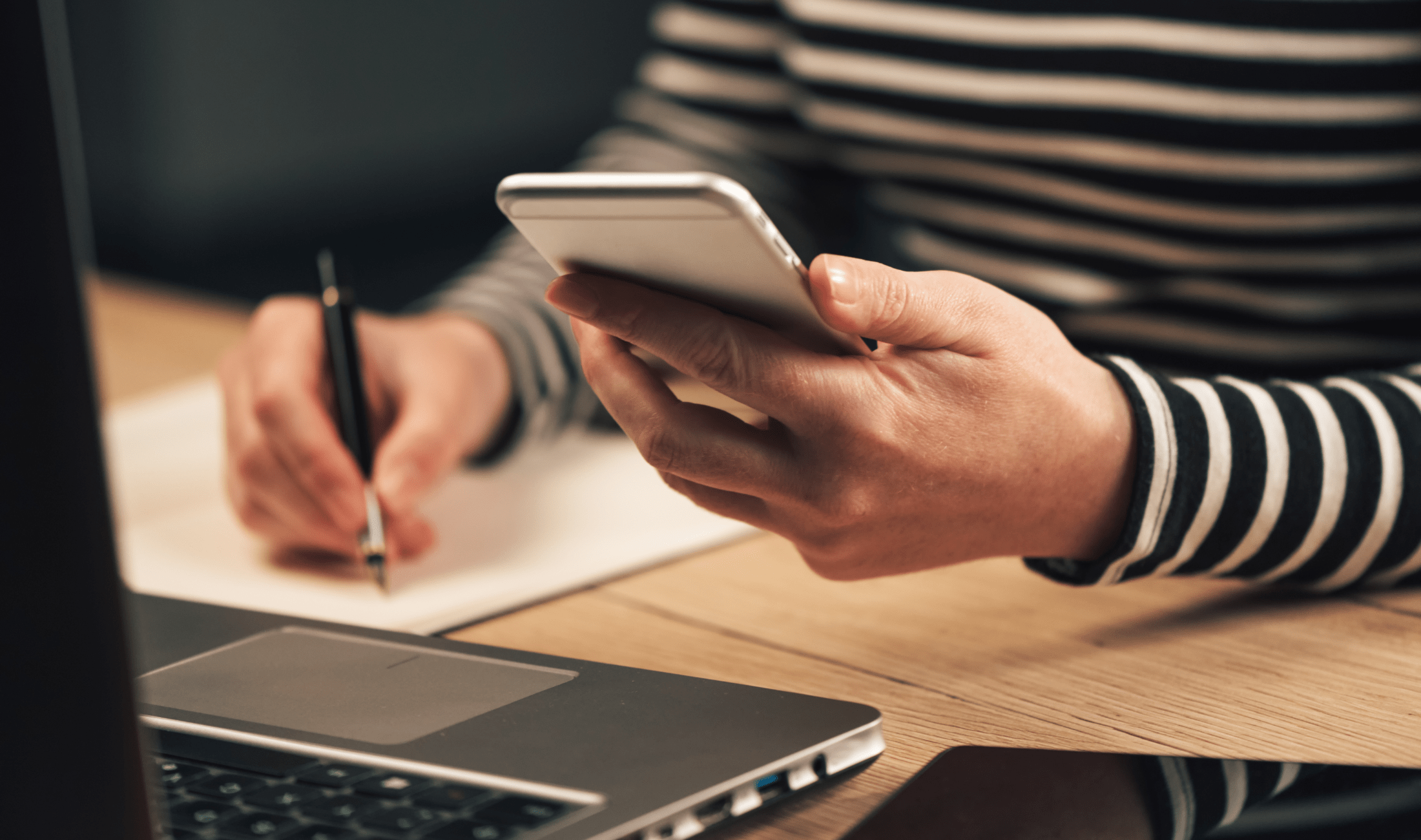 A woman sitting ay a table with her laptop, holding a phone in one hand, a pen in the other, about to copy something down.