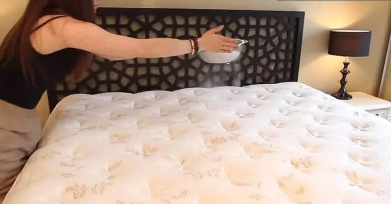 A woman holding a strainer sifting baking soda onto a bare mattress.