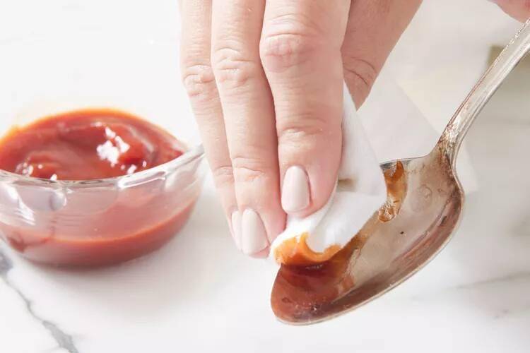 A hand using a napkin dipped in ketchup to clean a silver spoon.