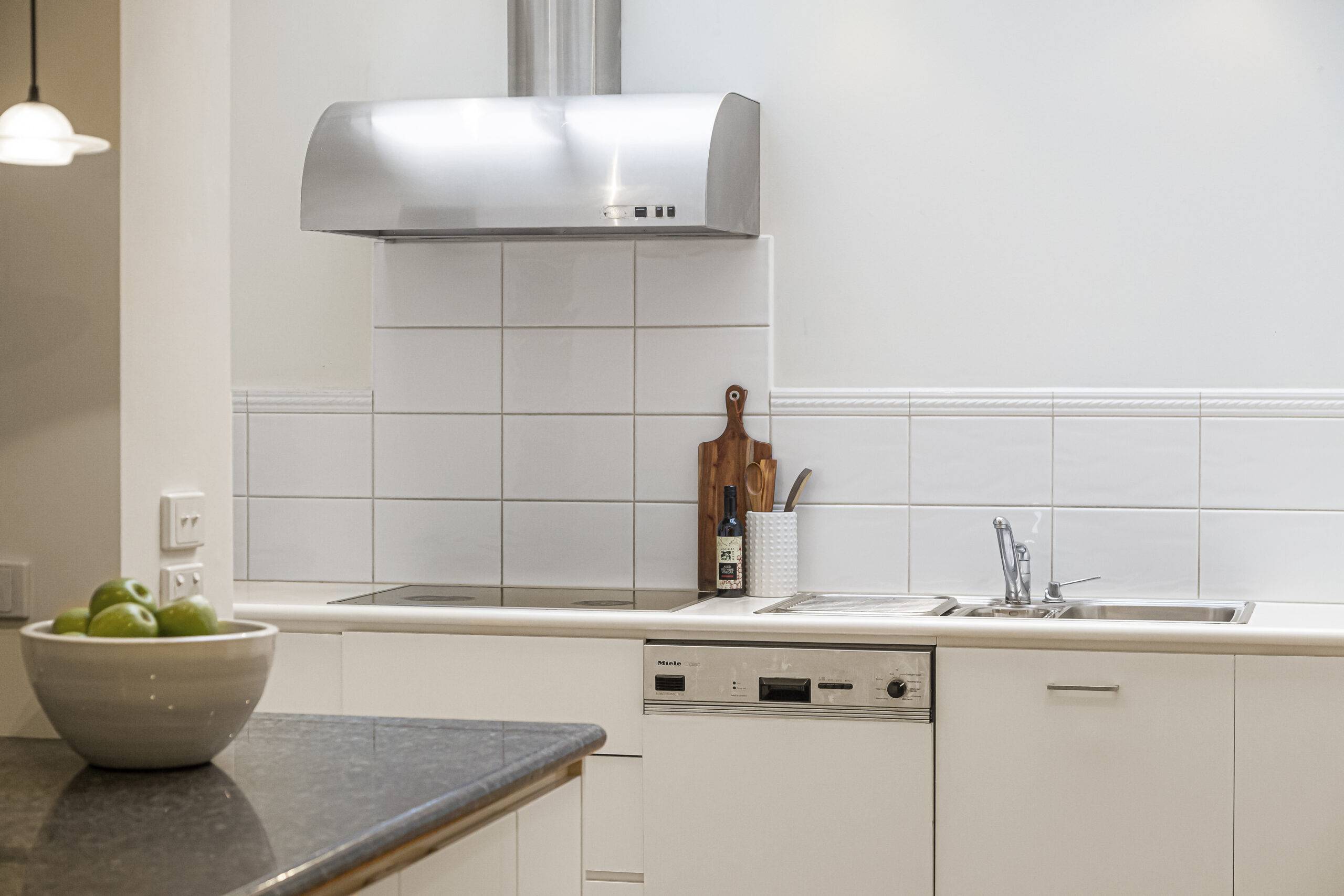 A white, clean kitchen, focus on a section of countertop with a sink, dishwasher, stovetop, and exhaust fan.