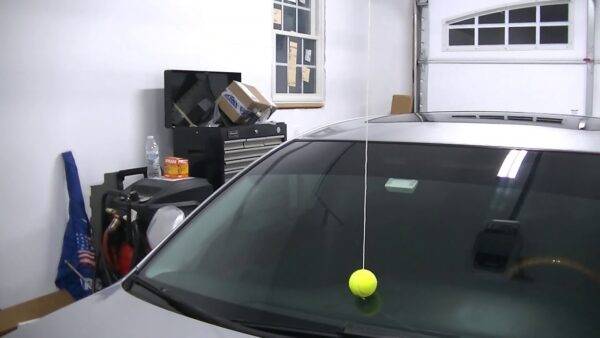 A tennis ball hanging from a string on the roof of a garage, the ball touching a car windshield.