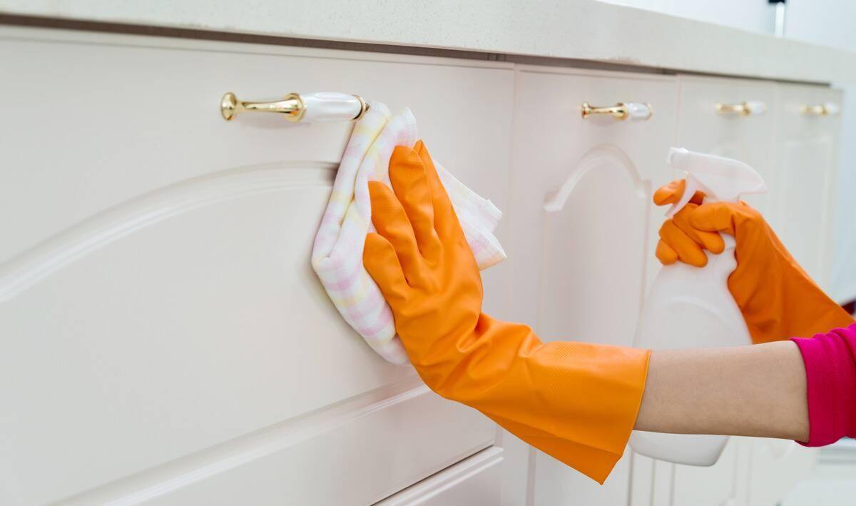 A close photo of a pair of arms in orange rubber gloves cleaning cabinets with a spray bottle and cloth.