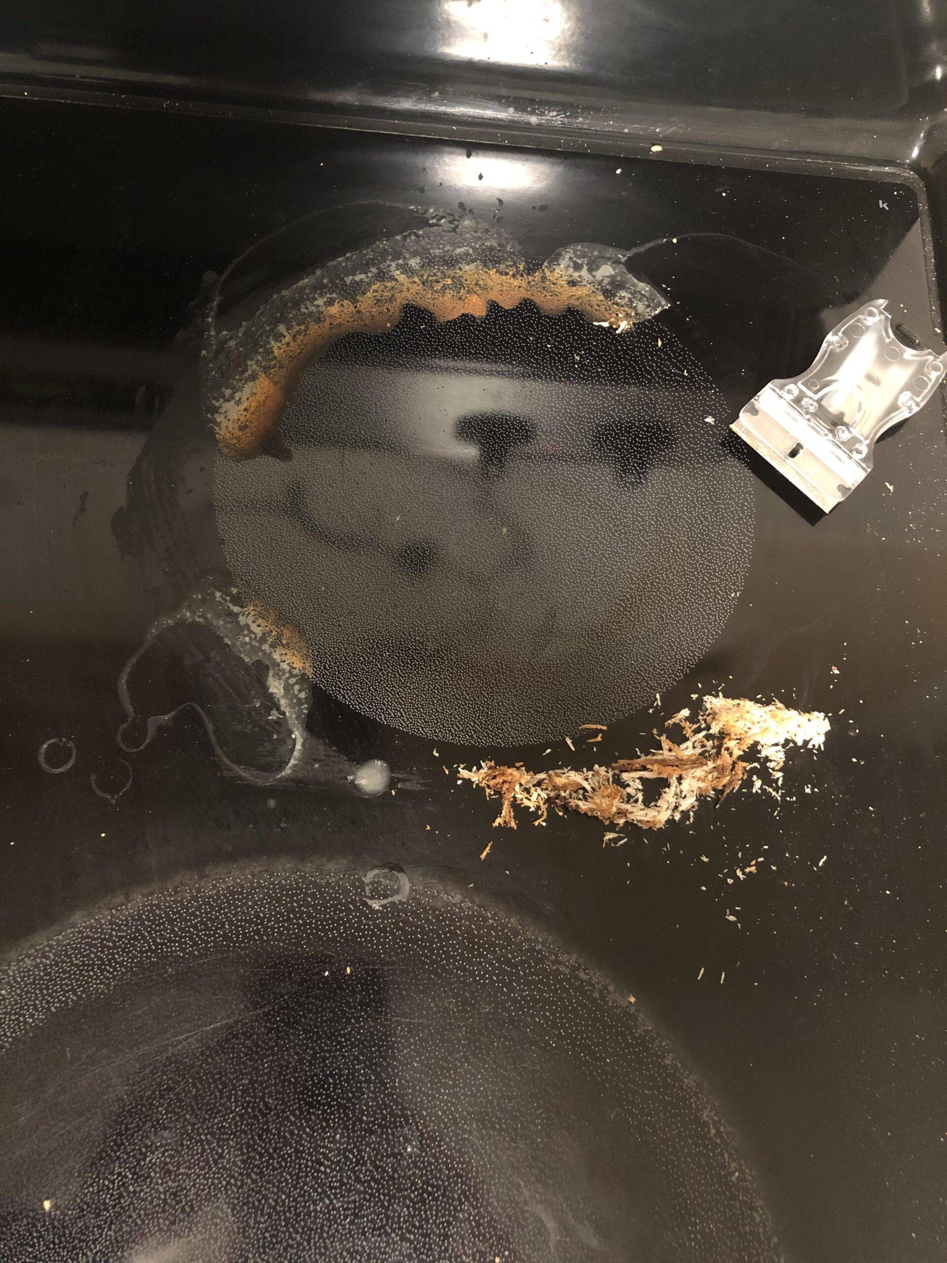A glass stovetop post-scraping, the mess lifted and piled together, the razor scraper placed near the top.