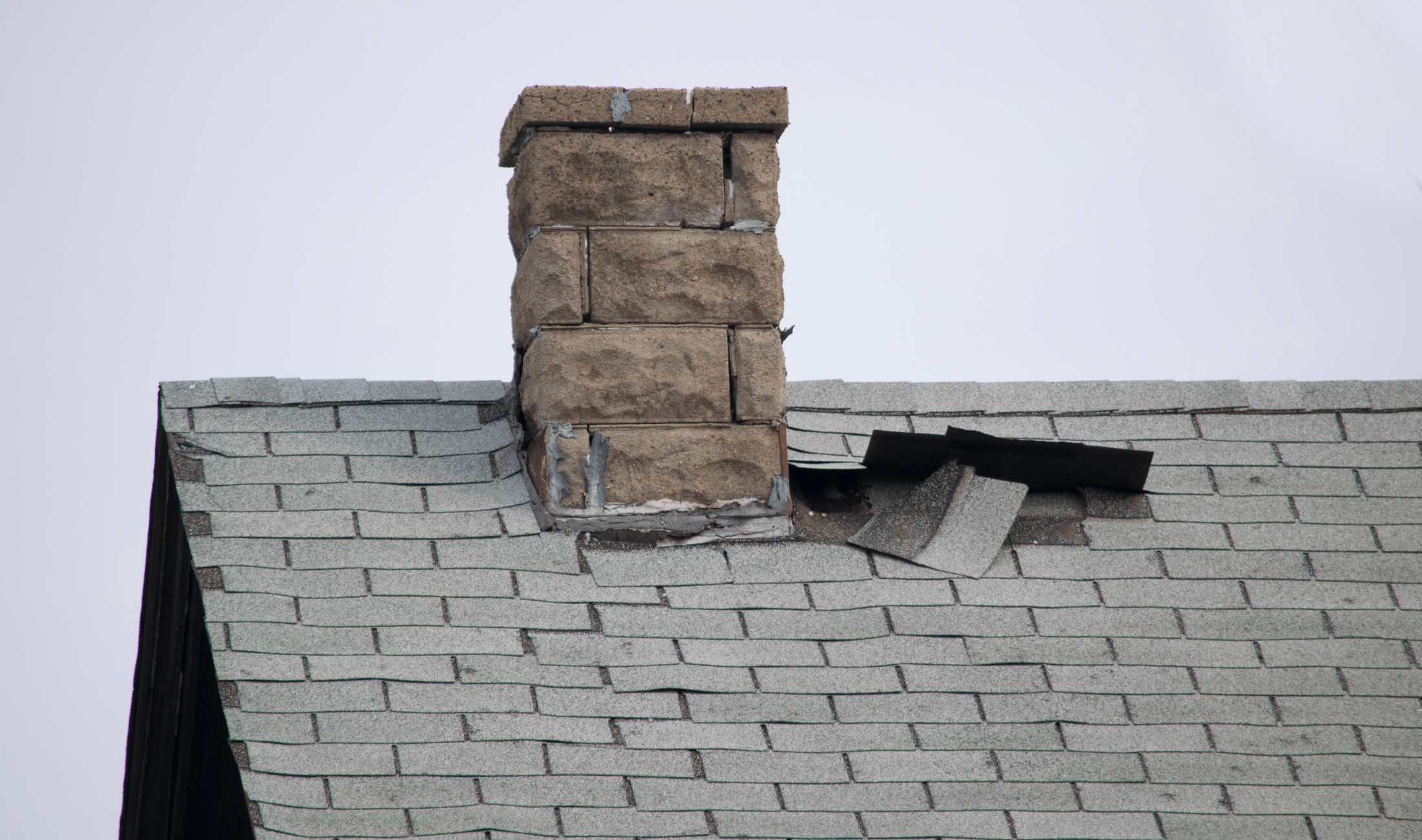 The roof of a home, some shingles misplaced next to the chimney.