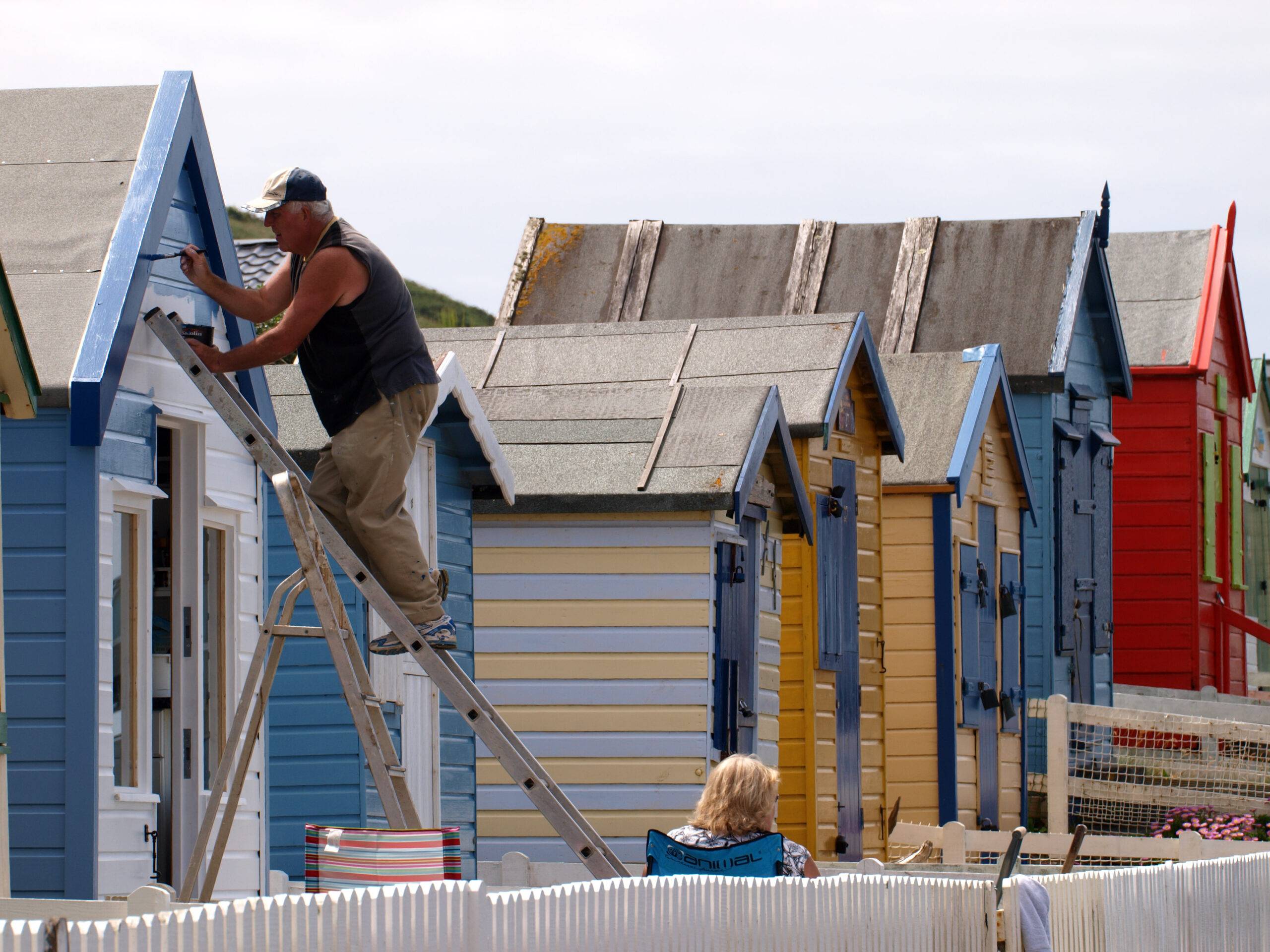 Old man on a ladder painting a beach hut 