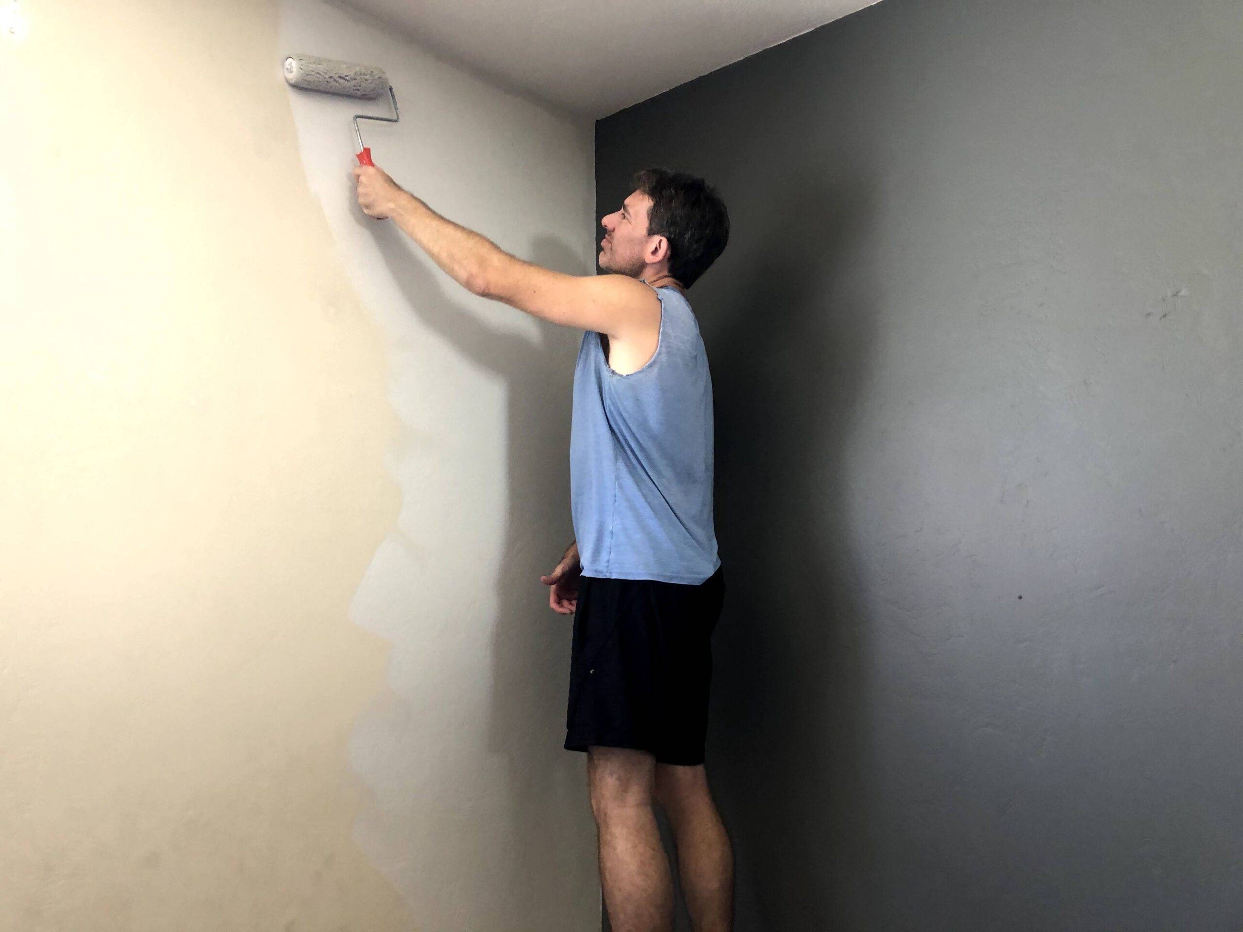 A man on a step stool using a paint roller to paint the upper corner of one wall.