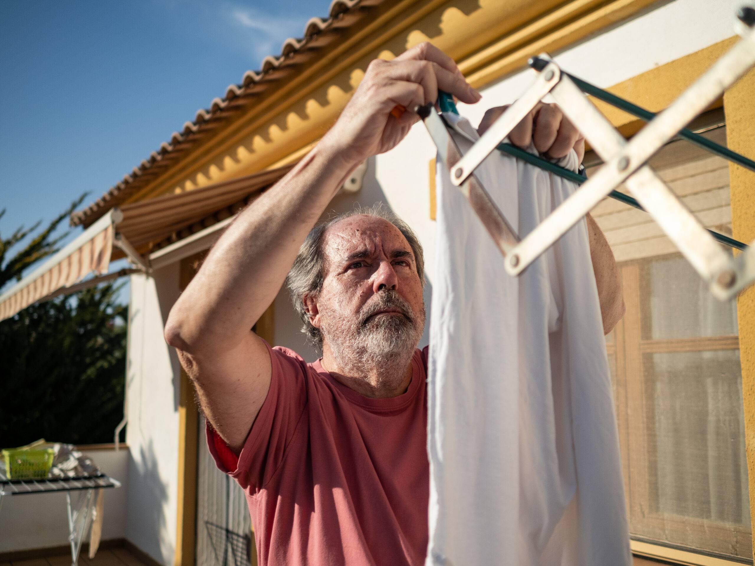 Senior man carefully hangs a white shirt on a clothesline outside a bright house. The warm sun emphasizes the tranquil atmosphere and simplicity of daily life.