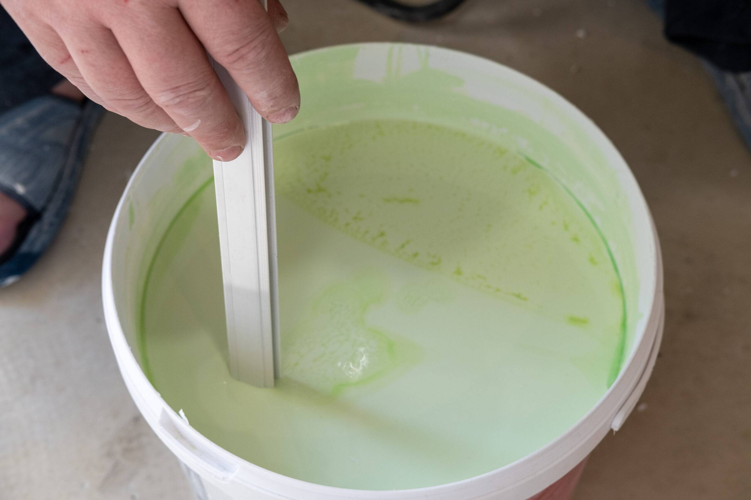 Worker's hands stir green paint before applying it, close-up. Bucket for mixing paints.